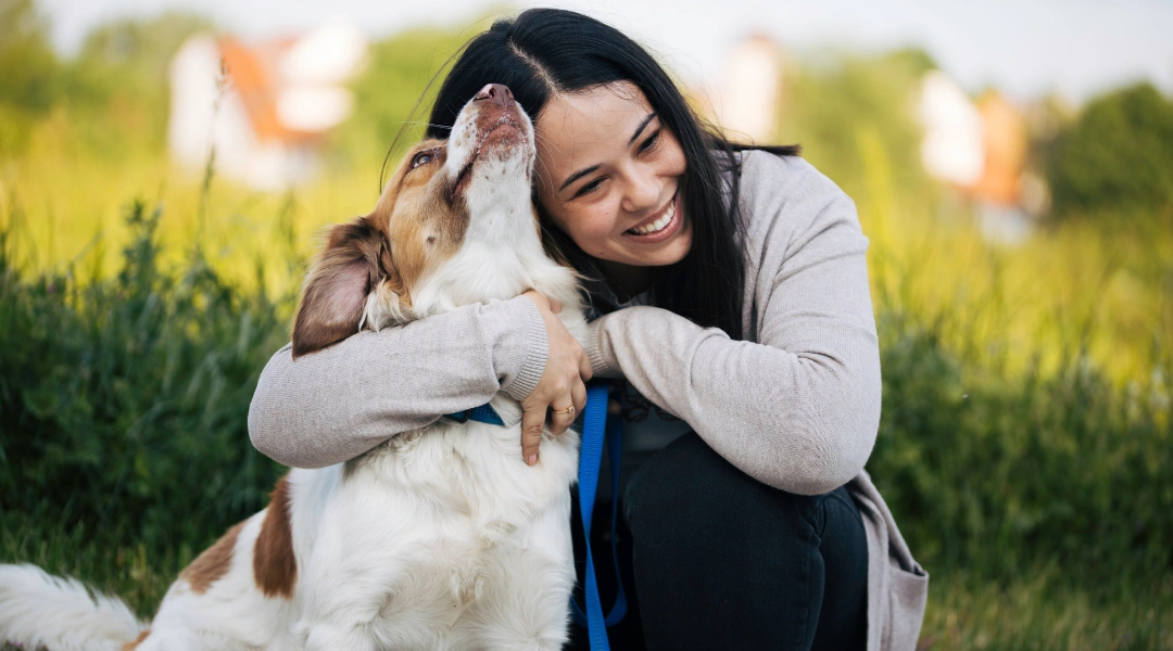 A woman sits on the grass, embracing her dog with a joyful expression | The Pet Tap | The Pet Tap Woman smiling while hugging a white and brown dog outdoors in a grassy field on a sunny day, showing affection.